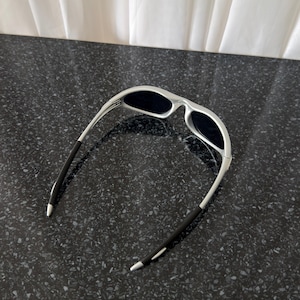 Puede incluir: Gafas de sol plateadas con lentes oscuras. Las gafas tienen un dise&ntilde;o curvo con patillas negras. Las lentes son oscuras y la montura es de color plata brillante. Las gafas de sol est&aacute;n sobre una superficie oscura y moteada.