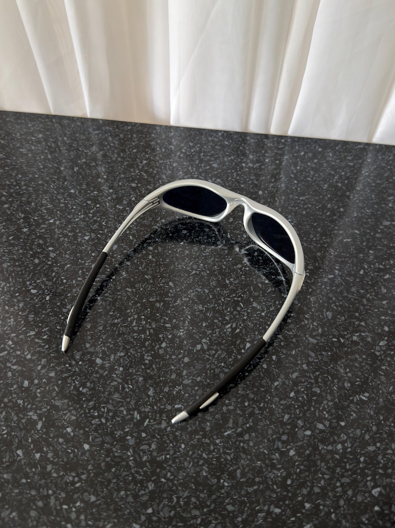 Puede incluir: Gafas de sol plateadas con lentes oscuras. Las gafas tienen un dise&ntilde;o curvo con patillas negras. Las lentes son oscuras y la montura es de color plata brillante. Las gafas de sol est&aacute;n sobre una superficie oscura y moteada.
