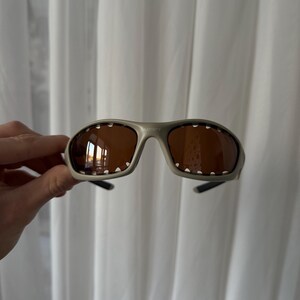May include: Gray sunglasses with brown lenses. The sunglasses have a sporty design with a curved frame and small white dots along the top edge of the lenses. The brand name "SATISFY" is printed on the frame.