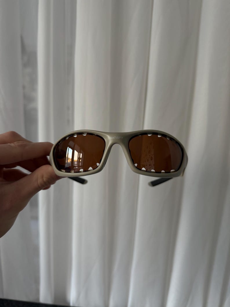 May include: Gray sunglasses with brown lenses. The sunglasses have a sporty design with a curved frame and small white dots along the top edge of the lenses. The brand name "SATISFY" is printed on the frame.