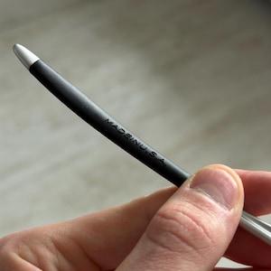 May include: A black stylus with a silver tip, held in a hand. The text "MADE IN U.S.A." is printed on the stylus. The stylus is curved and appears to be made of metal and rubber.