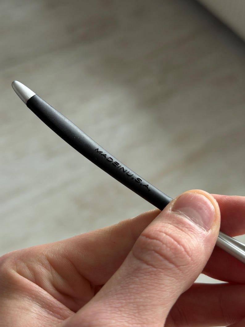 May include: A black stylus with a silver tip, held in a hand. The text "MADE IN U.S.A." is printed on the stylus. The stylus is curved and appears to be made of metal and rubber.
