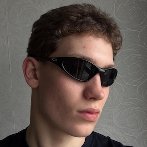 K&ouml;nnte beinhalten: Schwarze, ovale Sonnenbrille mit dunklen Gl&auml;sern. Die Sonnenbrille hat ein schlankes, sportliches Design mit einem geschwungenen Rahmen. Das Bild zeigt eine Nahaufnahme der Sonnenbrille.