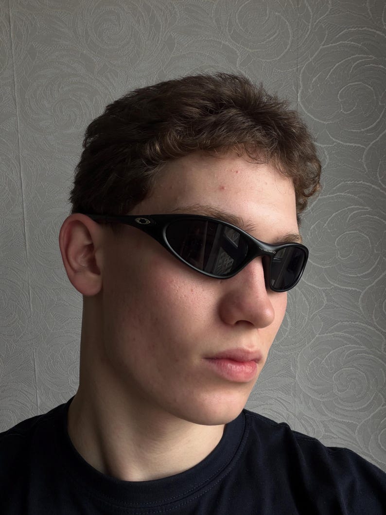 K&ouml;nnte beinhalten: Schwarze, ovale Sonnenbrille mit dunklen Gl&auml;sern. Die Sonnenbrille hat ein schlankes, sportliches Design mit einem geschwungenen Rahmen. Das Bild zeigt eine Nahaufnahme der Sonnenbrille.