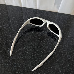 Puede incluir: Gafas de sol blancas con lentes negras. Las gafas tienen un dise&ntilde;o deportivo con una montura gruesa y patillas curvas. Las lentes son de forma ovalada y la montura es de color blanco liso.