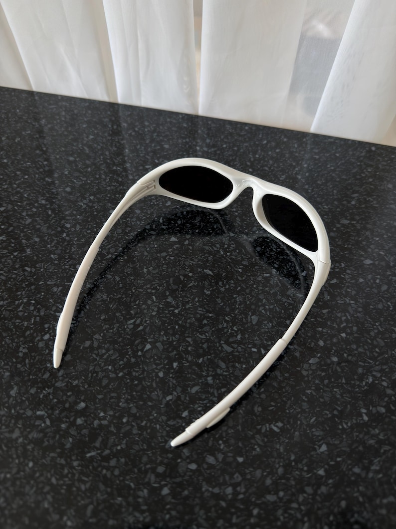 Puede incluir: Gafas de sol blancas con lentes negras. Las gafas tienen un dise&ntilde;o deportivo con una montura gruesa y patillas curvas. Las lentes son de forma ovalada y la montura es de color blanco liso.