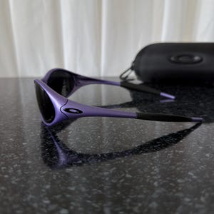 May include: Purple sunglasses with black lenses and a black case. The sunglasses have a sleek, modern design with a curved frame and a small logo on the side. The case is rectangular and has a zipper closure.