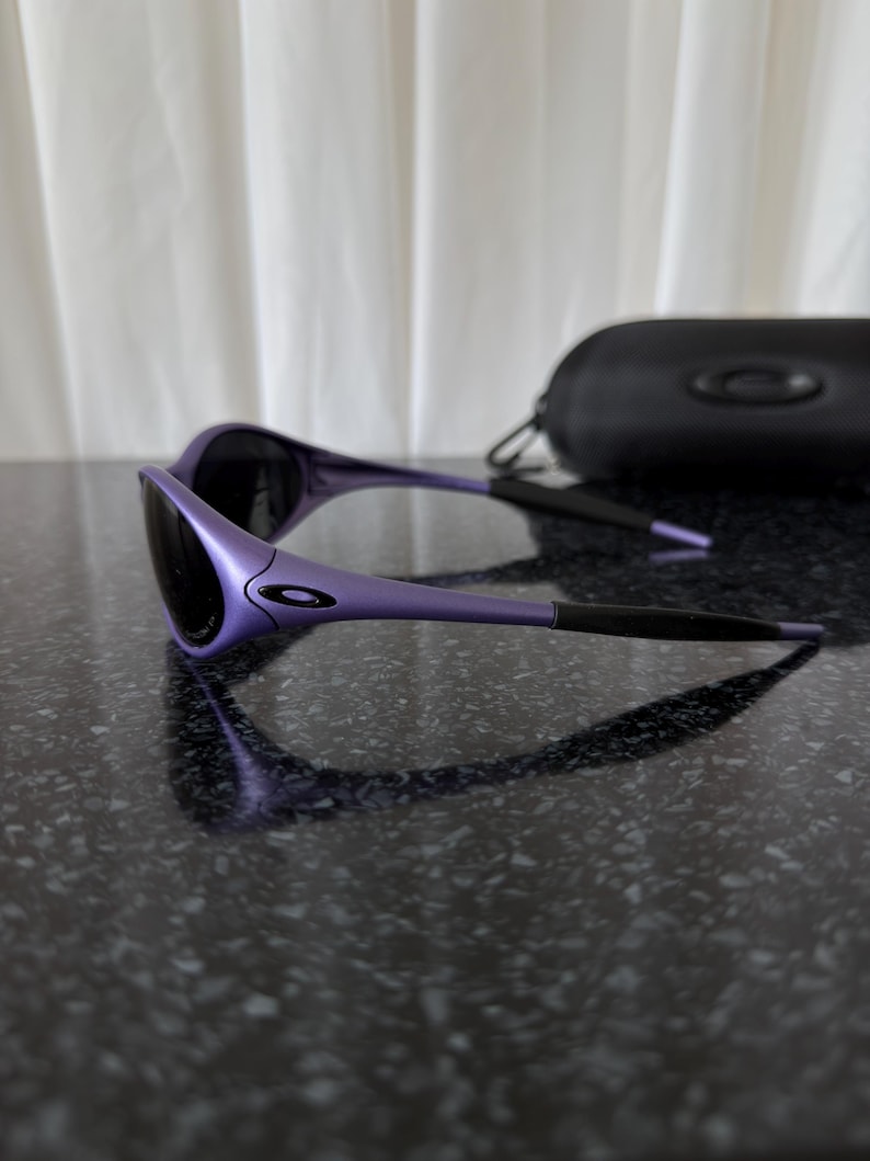 May include: Purple sunglasses with black lenses and a black case. The sunglasses have a sleek, modern design with a curved frame and a small logo on the side. The case is rectangular and has a zipper closure.