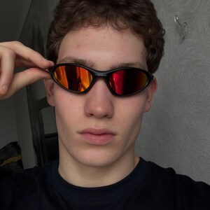May include: Black oval-shaped sunglasses with red-orange mirrored lenses. The sunglasses have a sleek, modern design and are held by a person. The frame is a matte black color.