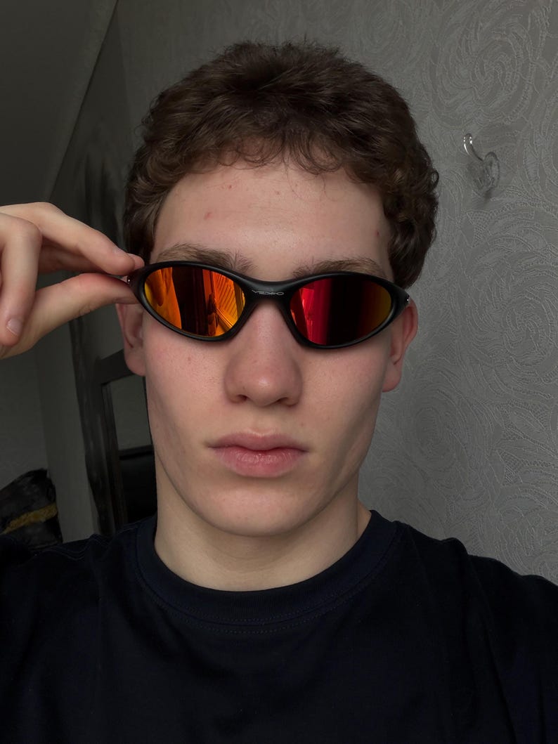 May include: Black oval-shaped sunglasses with red-orange mirrored lenses. The sunglasses have a sleek, modern design and are held by a person. The frame is a matte black color.