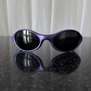 May include: Purple sunglasses with black lenses. The sunglasses have a curved, oval shape and a glossy finish. The lenses reflect the surrounding environment. The sunglasses are resting on a dark, reflective surface.