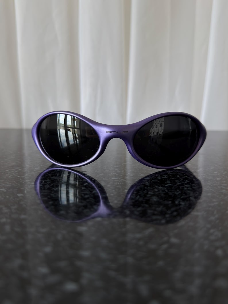 May include: Purple sunglasses with black lenses. The sunglasses have a curved, oval shape and a glossy finish. The lenses reflect the surrounding environment. The sunglasses are resting on a dark, reflective surface.