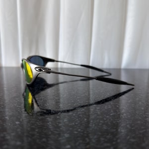 May include: Sleek, modern sunglasses with a silver frame and black temples. The lenses have a mirrored finish, reflecting yellow and blue hues. The sunglasses are resting on a dark, glossy surface, with a white curtain in the background.