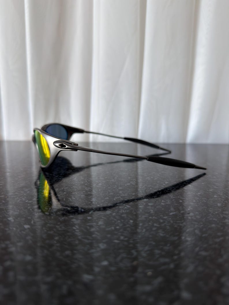 May include: Sleek, modern sunglasses with a silver frame and black temples. The lenses have a mirrored finish, reflecting yellow and blue hues. The sunglasses are resting on a dark, glossy surface, with a white curtain in the background.