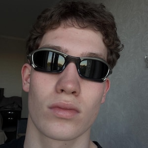 May include: Sport sunglasses with a silver frame and dark lenses. The sunglasses have a wraparound design and are worn by a person. The lenses reflect the surrounding environment.