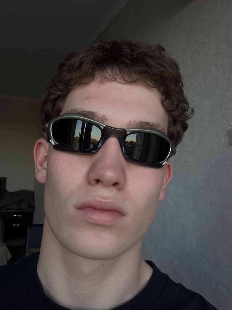 May include: Sport sunglasses with a silver frame and dark lenses. The sunglasses have a wraparound design and are worn by a person. The lenses reflect the surrounding environment.