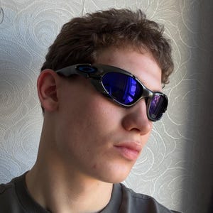 Op de afbeelding: Zwarte en zilveren zonnebril met blauwe reflecterende glazen. De zonnebril heeft een gebogen, omsluitend ontwerp. Het montuur is donker, met een subtiele metallic glans. De glazen zijn helderblauw.
