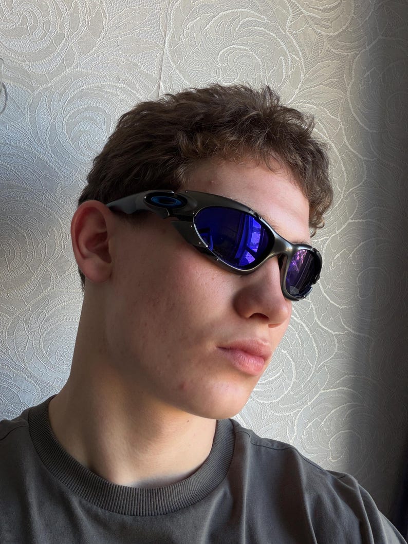 Op de afbeelding: Zwarte en zilveren zonnebril met blauwe reflecterende glazen. De zonnebril heeft een gebogen, omsluitend ontwerp. Het montuur is donker, met een subtiele metallic glans. De glazen zijn helderblauw.
