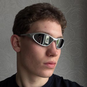 May include: Silver-framed sunglasses with a curved design and mirrored lenses. The sunglasses have a sleek, modern aesthetic. The lenses reflect the surrounding environment. The frame is a light silver color.