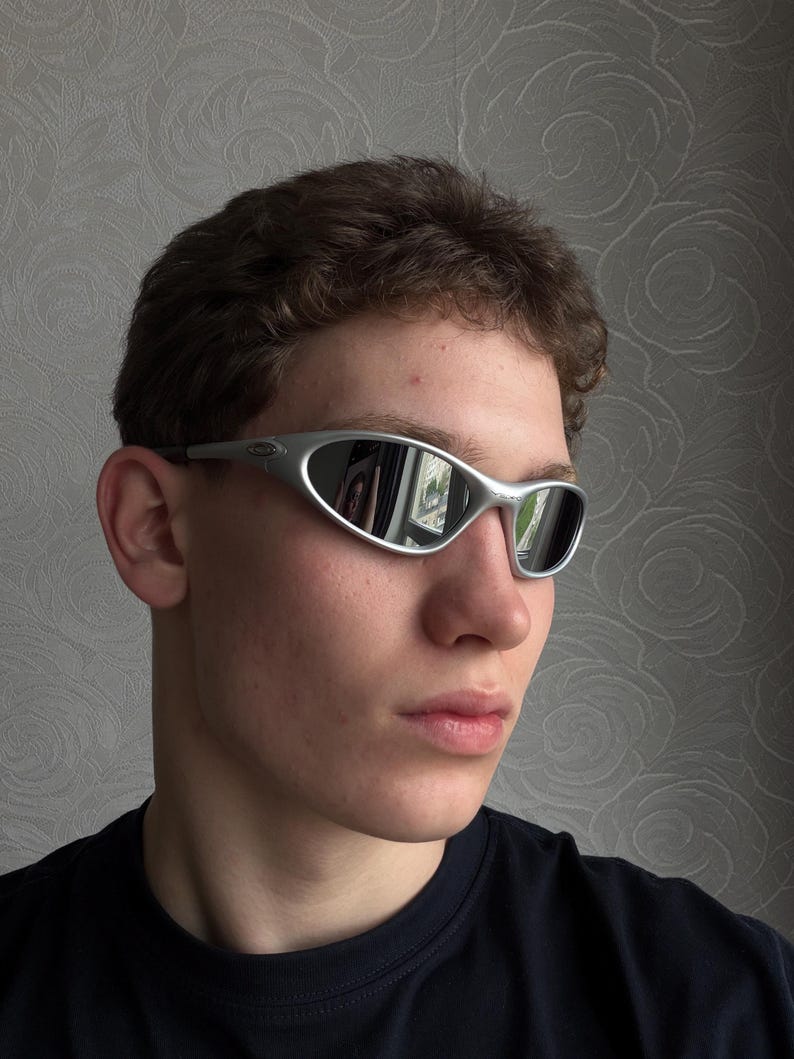 May include: Silver-framed sunglasses with a curved design and mirrored lenses. The sunglasses have a sleek, modern aesthetic. The lenses reflect the surrounding environment. The frame is a light silver color.