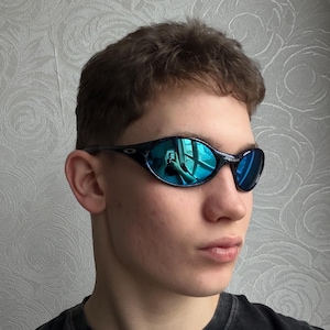 K&ouml;nnte beinhalten: Schwarz-blaue Sonnenbrille mit reflektierenden blauen Gl&auml;sern. Die Sonnenbrille hat einen dunklen Rahmen und ein geschwungenes Design. Die Gl&auml;ser reflektieren eine leuchtend blaue Farbe. Die Sonnenbrille ist sportlich.