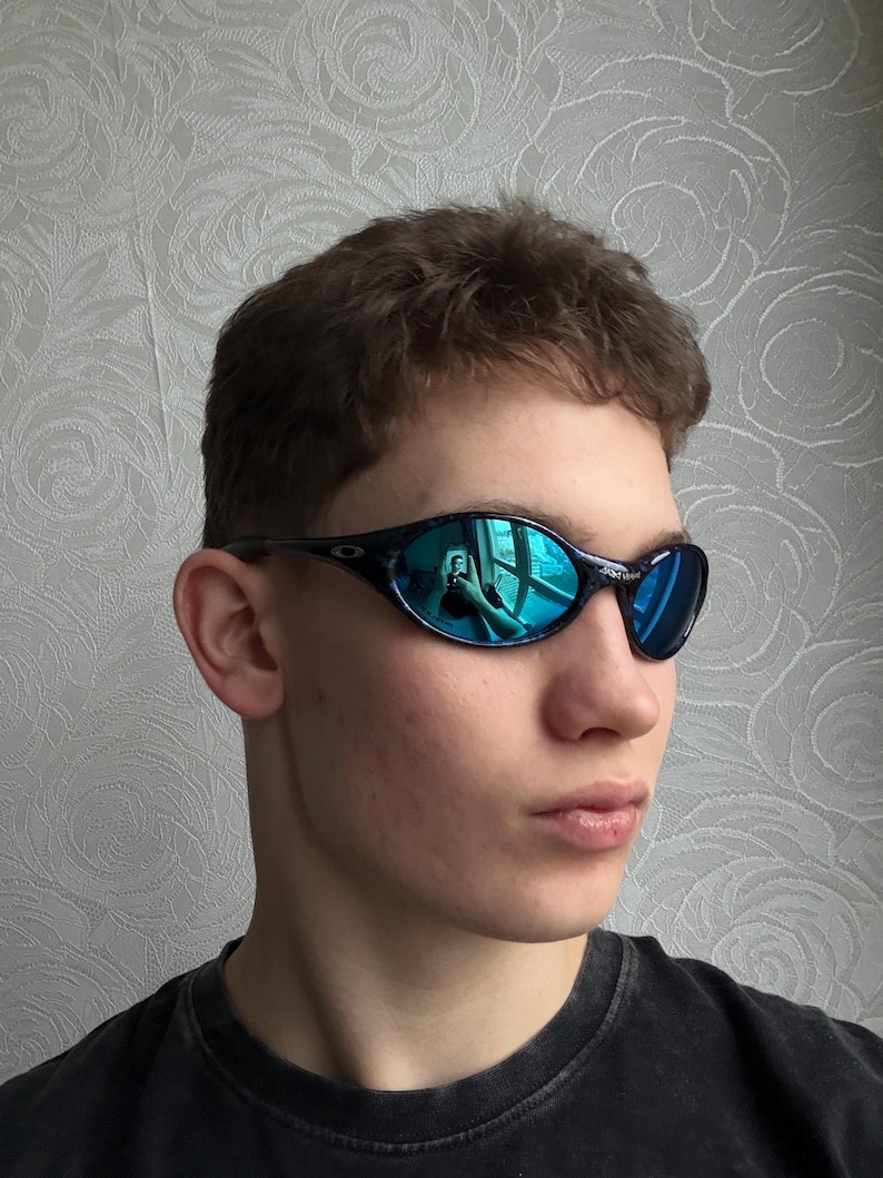K&ouml;nnte beinhalten: Schwarz-blaue Sonnenbrille mit reflektierenden blauen Gl&auml;sern. Die Sonnenbrille hat einen dunklen Rahmen und ein geschwungenes Design. Die Gl&auml;ser reflektieren eine leuchtend blaue Farbe. Die Sonnenbrille ist sportlich.