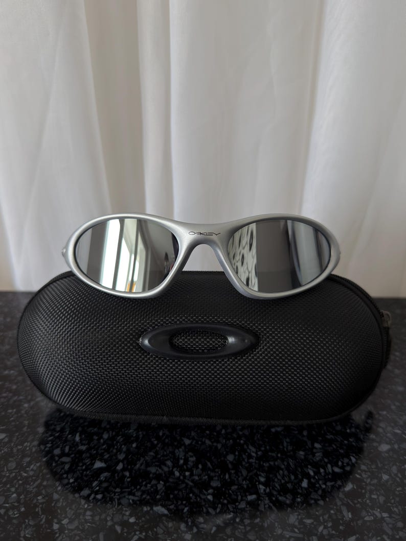 May include: Silver-framed sunglasses with reflective lenses rest on a black, textured case. The sunglasses have a sleek, modern design. The case has a rounded shape and a subtle logo.