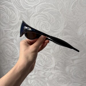 K&ouml;nnte beinhalten: Schwarze und blaue Sonnenbrille mit gebogenem Rahmen und get&ouml;nten Gl&auml;sern. Die Sonnenbrille hat ein schlankes, modernes Design mit dunklem Rahmen und blau get&ouml;nten Gl&auml;sern. Die Sonnenbrille wird von einer Hand gehalten.