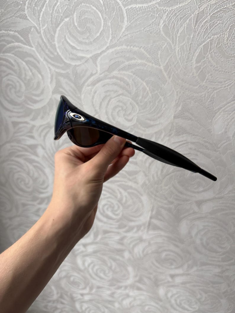 K&ouml;nnte beinhalten: Schwarze und blaue Sonnenbrille mit gebogenem Rahmen und get&ouml;nten Gl&auml;sern. Die Sonnenbrille hat ein schlankes, modernes Design mit dunklem Rahmen und blau get&ouml;nten Gl&auml;sern. Die Sonnenbrille wird von einer Hand gehalten.