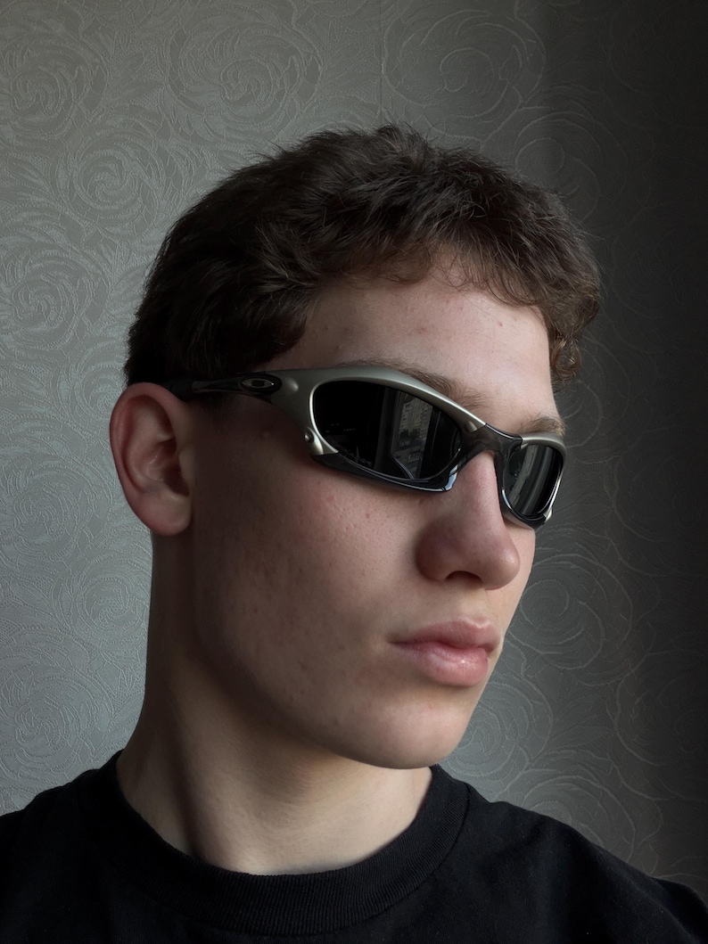 May include: Silver-framed sunglasses with dark lenses. The sunglasses have a sleek, modern design with a curved frame and a metallic finish. The lenses are reflective, and the overall style is sporty and stylish.