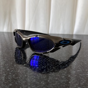 Op de afbeelding: Sportzonnebril met een metallic grijs montuur en blauwe glazen. De zonnebril heeft een strak, modern design met een gebogen vorm en een klein blauw logo op de poot. De glazen reflecteren een helderblauw licht.