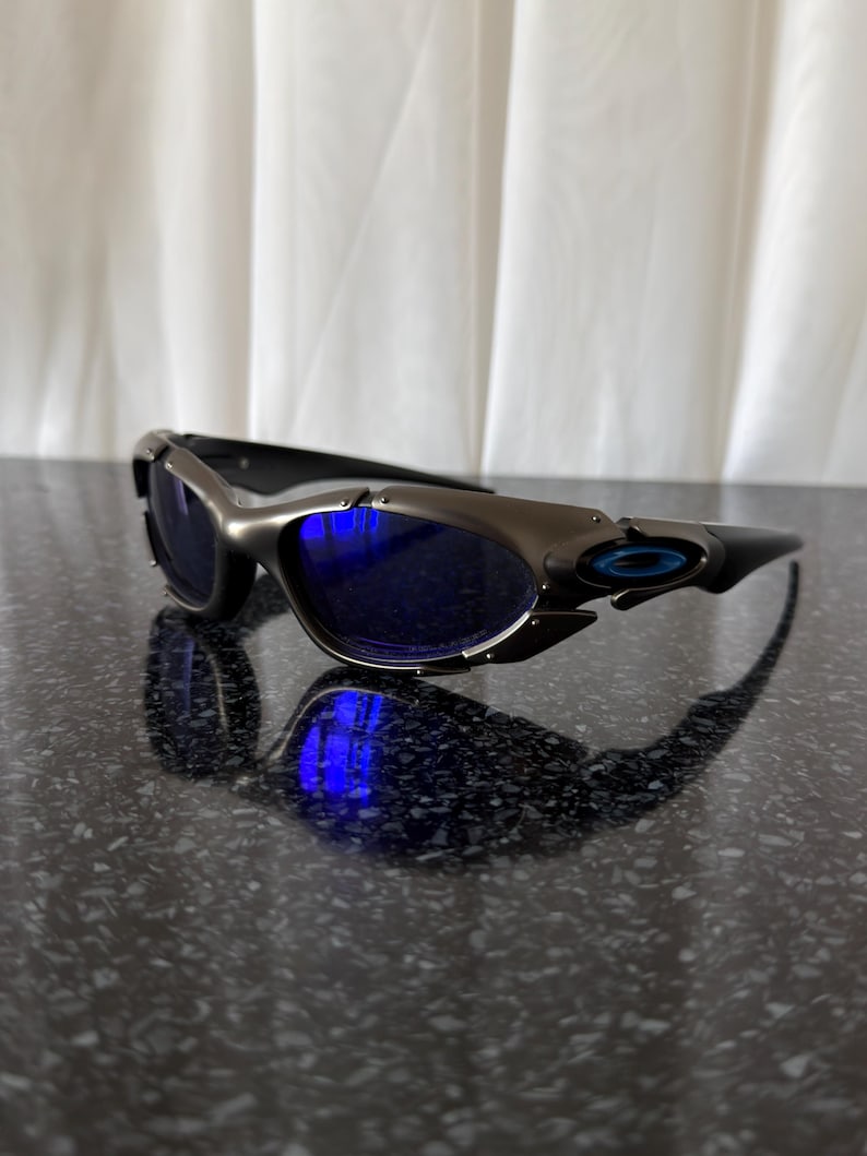 Op de afbeelding: Sportzonnebril met een metallic grijs montuur en blauwe glazen. De zonnebril heeft een strak, modern design met een gebogen vorm en een klein blauw logo op de poot. De glazen reflecteren een helderblauw licht.