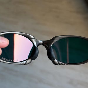 May include: Close-up of a pair of sunglasses with a silver frame and polarized lenses. The lenses have a gradient of pink and green hues. The nose pads are black, and the frame has a sleek, modern design. The text "POLARIZED" is visible on the frame.