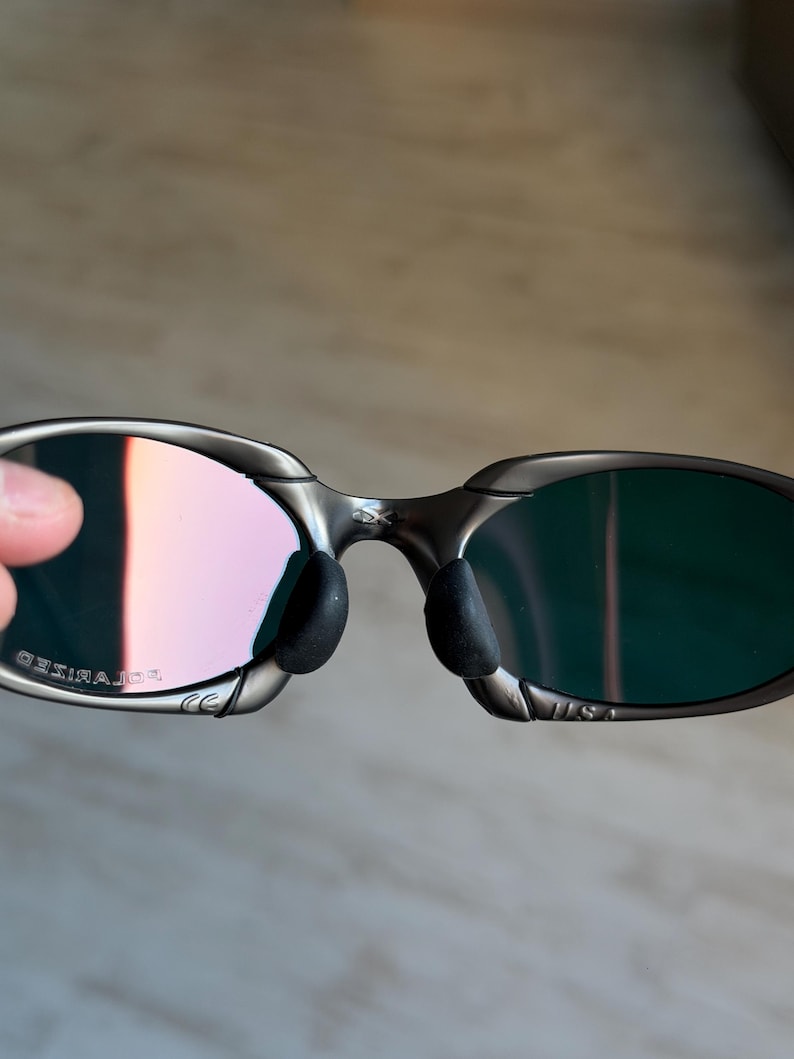 May include: Close-up of a pair of sunglasses with a silver frame and polarized lenses. The lenses have a gradient of pink and green hues. The nose pads are black, and the frame has a sleek, modern design. The text "POLARIZED" is visible on the frame.