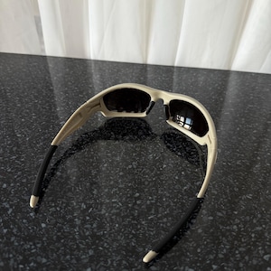 May include: Off-white sunglasses with black lenses and black temple tips. The sunglasses have a curved design and are set against a dark speckled surface.