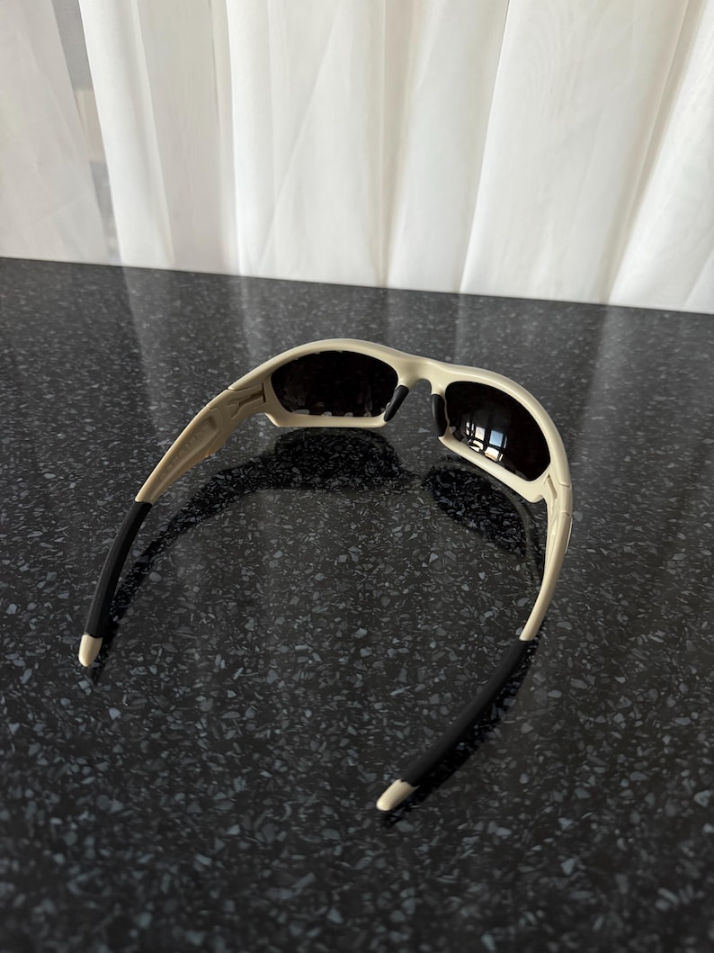 May include: Off-white sunglasses with black lenses and black temple tips. The sunglasses have a curved design and are set against a dark speckled surface.