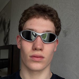May include: Silver-framed sunglasses with oval lenses. The sunglasses have a sleek, modern design. The lenses are dark, reflecting the surrounding environment. The frame is a shiny silver color. The sunglasses are a stylish accessory.