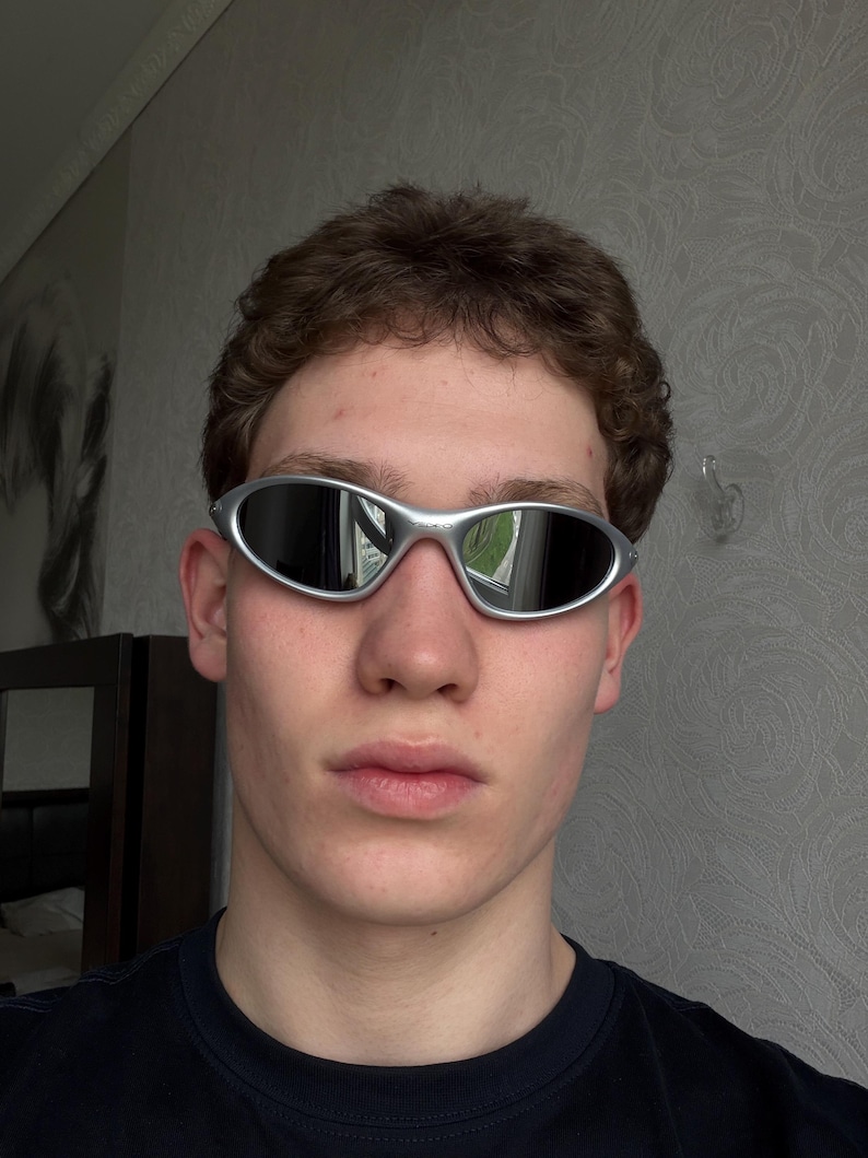 May include: Silver-framed sunglasses with oval lenses. The sunglasses have a sleek, modern design. The lenses are dark, reflecting the surrounding environment. The frame is a shiny silver color. The sunglasses are a stylish accessory.