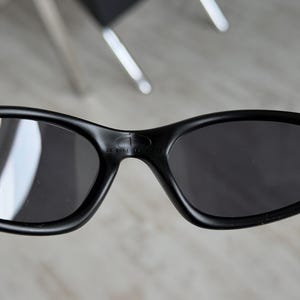 K&ouml;nnte beinhalten: Nahaufnahme einer schwarzen Sonnenbrille mit dunklen Gl&auml;sern. Die Sonnenbrille hat einen geschwungenen Rahmen und der Markenname "MINUS" ist auf dem Steg sichtbar. Die Gl&auml;ser sind dunkel und reflektieren die Umgebung.