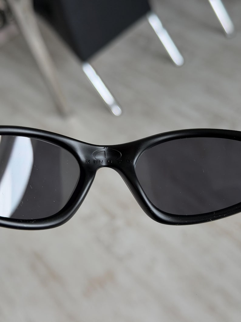 K&ouml;nnte beinhalten: Nahaufnahme einer schwarzen Sonnenbrille mit dunklen Gl&auml;sern. Die Sonnenbrille hat einen geschwungenen Rahmen und der Markenname "MINUS" ist auf dem Steg sichtbar. Die Gl&auml;ser sind dunkel und reflektieren die Umgebung.