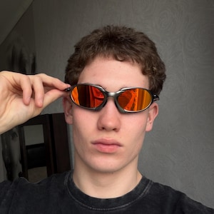 May include: Oval-shaped sunglasses with a silver frame and orange-tinted lenses. The sunglasses are held up by a hand. The person is wearing a black t-shirt.