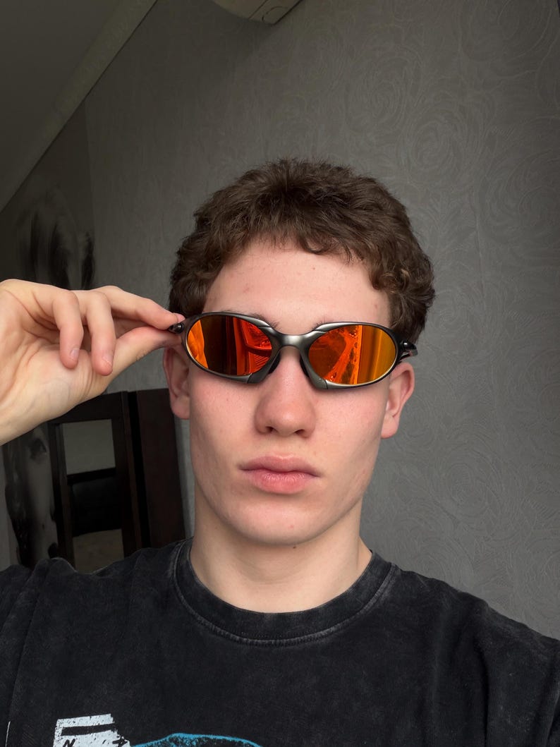 May include: Oval-shaped sunglasses with a silver frame and orange-tinted lenses. The sunglasses are held up by a hand. The person is wearing a black t-shirt.