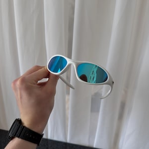Puede incluir: Gafas de sol blancas con lentes azules espejadas. Las gafas tienen una montura y patillas curvas. Las lentes reflejan el entorno. Las gafas se sostienen frente a una cortina blanca.