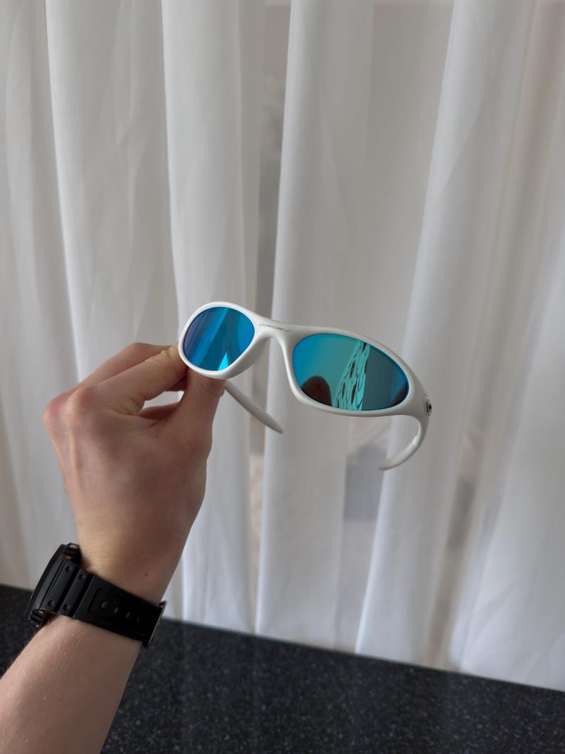 Puede incluir: Gafas de sol blancas con lentes azules espejadas. Las gafas tienen una montura y patillas curvas. Las lentes reflejan el entorno. Las gafas se sostienen frente a una cortina blanca.