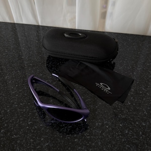 May include: Purple sunglasses with a black case and a black cleaning cloth. The sunglasses have a sleek, modern design with dark lenses. The case is black and has the brand logo. The cleaning cloth has the brand name and logo.