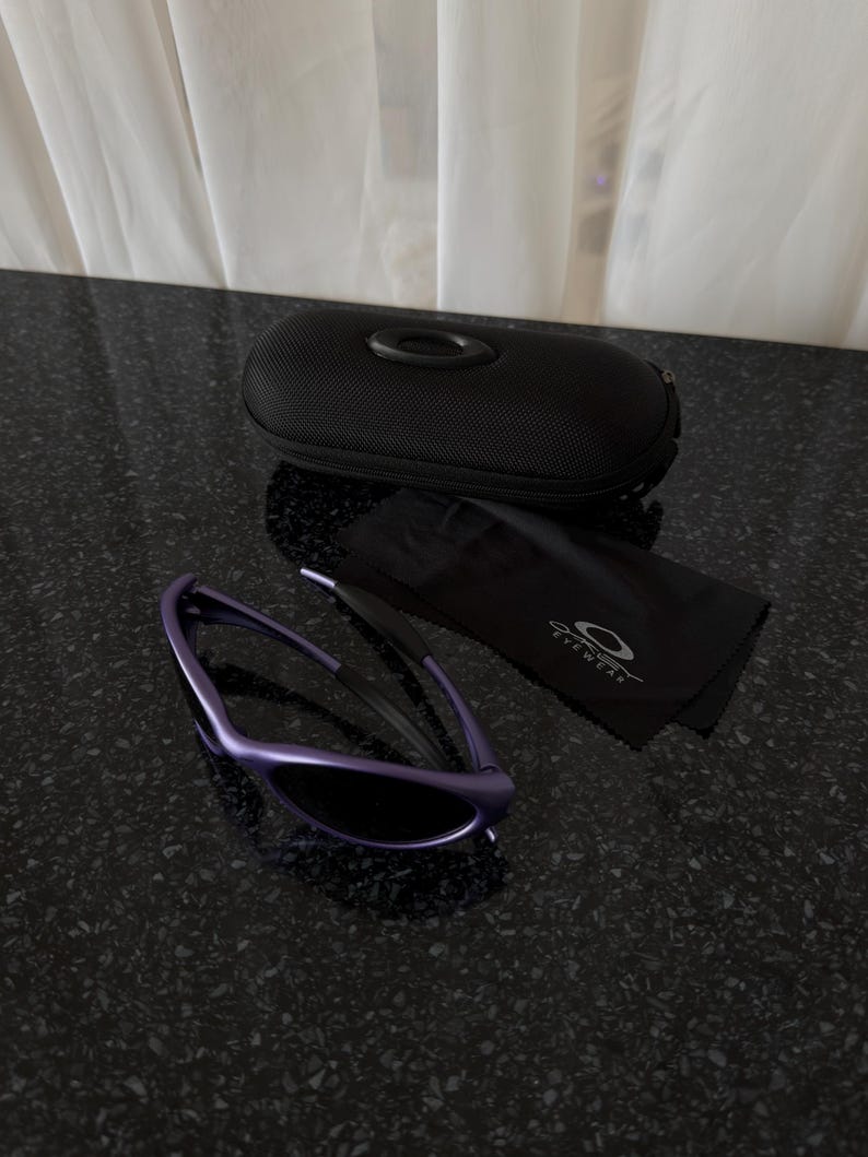 May include: Purple sunglasses with a black case and a black cleaning cloth. The sunglasses have a sleek, modern design with dark lenses. The case is black and has the brand logo. The cleaning cloth has the brand name and logo.