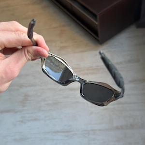 May include: Silver and black sunglasses with a rectangular frame and dark lenses. The temples are black and curved, with a silver accent near the lenses. The sunglasses are held by a hand.