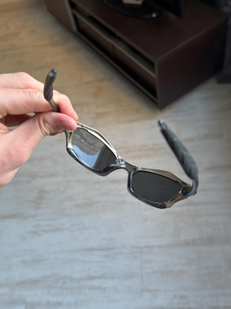May include: Silver and black sunglasses with a rectangular frame and dark lenses. The temples are black and curved, with a silver accent near the lenses. The sunglasses are held by a hand.