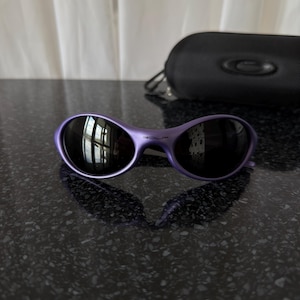 May include: Purple sunglasses with dark lenses and a matching case. The sunglasses have a curved frame and a sleek, modern design. The case is black and features a logo.