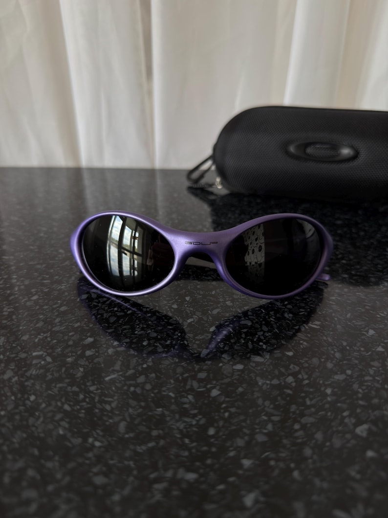 May include: Purple sunglasses with dark lenses and a matching case. The sunglasses have a curved frame and a sleek, modern design. The case is black and features a logo.